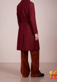 Persona de pie de espaldas, vistiendo un largo abrigo burdeos y pantalones de pana anchos de color marrón, frente a un fondo simple.