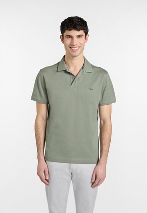 Giovane uomo sorridente, che indossa una polo verde chiaro e pantaloni grigio chiaro, in piedi contro uno sfondo bianco uniforme.