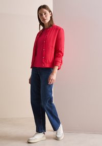 Giacca bomber rossa trapuntata con chiusure a pressione, indossata sopra un top rosa e abbinata a jeans blu e sneakers bianche. Tessuto morbido, vestibilità casual.