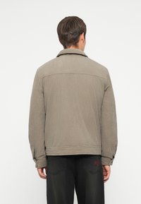 Khaki corduroy-takki, jossa on suuri kaulus, näkyvä textura ja röyhelö, yhdistettynä mustiin housuihin, joissa on punainen logo takataskussa.