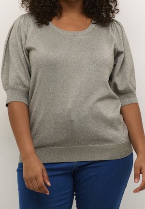 Kvinde iført en sølvfarvet kortærmet sweater med puffede skuldre og blå jeans, stående foran en ensfarvet lys baggrund.