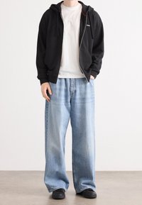 Černá mikina na zip s kapucí a šňůrkami přes bílé tričko, doplněná volnými světle modrými džínami a černými teniskami.