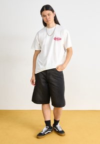Camiseta blanca con logo rojo de "Carhartt"; pantalones cortos de mezclilla negros; zapatillas Vans negras con acentos blancos; cadena plateada; pose de pie.