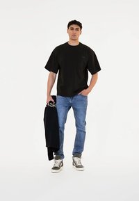 Černé tričko s krátkým rukávem, volný střih, spárované s světle modrými džíny. Model drží tmavý svetr s bílým logem a má na sobě šedé tenisky.