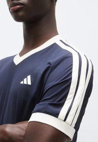 Αθλητική μπλούζα σε ναυτικό μπλε με λευκή επένδυση, ντεκολτέ σε σχήμα V και τρεις οριζόντιες ρίγες στα μανίκια. Διαθέτει λευκό λογότυπο στο στήθος.