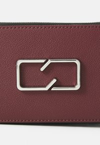 Portefeuille en cuir texturé bordeaux avec un fermoir rectangulaire en métal argenté. Il présente une finition lisse et des coutures visibles sur les bords.