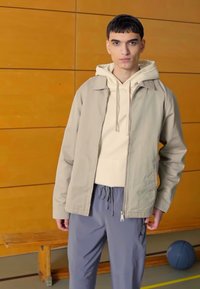 Beige letvægtsjakke over en creme hoodie, parret med grå træningsbukser med snor i taljen og lynlåslommer. Trævæg.