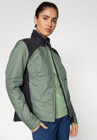 Veste matelassée avec un devant vert et des épaules noires, dotée d'un col montant, d'une fermeture éclair et d'un tissu texturé pour l'isolation.