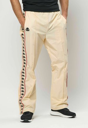 Beige Sporthose mit schwarzem Logo, roten Seitenstreifen, elastischem Bund, zwei Seitentaschen und weiter Passform, kombiniert mit schwarzen Turnschuhen.