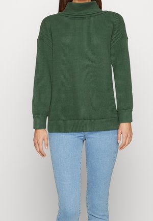 Femme portant un col roulé côtelé vert foncé et un jean bleu clair, debout devant un fond blanc uni.