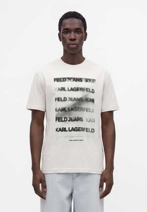 Νέος άνδρας με λευκό T-shirt με επαναλαμβανόμενο κείμενο "KARL LAGERFELD" και "FELD JEANS" σε θολό στυλ, ανοιχτόχρωμα τζιν, σε λευκό φόντο.