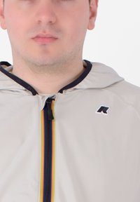Giacca zip con cappuccio di un beige chiaro, con bordo blu navy e una striscia verticale rossa, gialla e blu sul davanti. Logo sul petto.