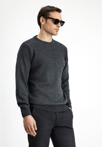 Suéter gris de punto con patrón texturizado en el pecho y puños acanalados. Combinado con pantalones oscuros, modelo lleva gafas de sol negras.
