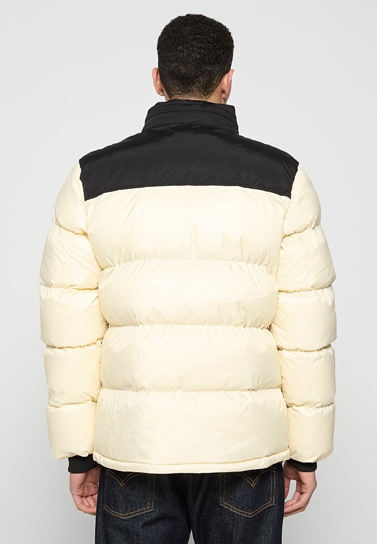 Mann in einer cremefarbenen und schwarzen Steppjacke, der sich abwendet, kombiniert mit dunkelblauen Jeans, vor einem schlichten hellen Hintergrund.