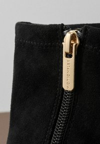 Bottine noire en suédine avec une fermeture éclair en métal doré portant le logo gravé "MOTIONFLEX". La texture est douce et lisse.