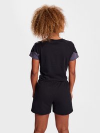 Woman with curly hair wearing a black t-shirt with gray sleeves and black shorts, standing facing away against a plain background.