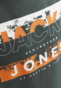 Temnozeleni T-shirt z bold belim in oranžnim grafičnim besedilom. Besedilo vključuje "JACK & JONES" z obrabljenim vzorcem in številčnimi podrobnostmi.