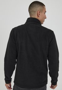 Chaqueta de forro polar negra con un cuello alto, tejido texturizado y mangas largas. Sin logotipos ni patrones visibles. Se muestra la vista trasera.