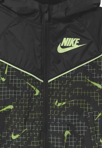 Černá Nike bunda s kapucí, ozdobená geometrickým zeleným mřížkovým vzorem. Doplněná o zelené logo a kontrastní bílé pruhy. Zapínání na zip na přední straně.