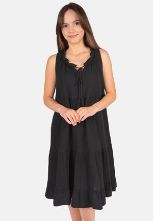 Femme aux longs cheveux bruns portant une robe noire sans manches à volants avec un nœud à l'encolure, debout devant un fond uni.
