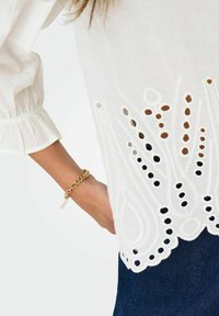 Weiße Bluse mit bestickten Ausschnitten am Saum. Sie hat einen lockeren Schnitt, puffige Ärmel und ein strukturiertes Design. Kombiniert mit dunklen Jeans.