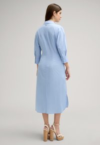 Helles blaues, langärmeliges Kleid mit einem Knopfdetail auf der Rückseite, geknöpften Manschetten, seitlichen Schlitzen und einer geraden Silhouette, kombiniert mit Absatzsandalen.
