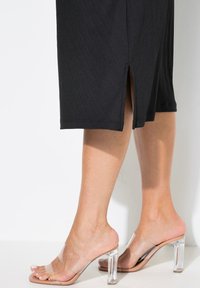 Transparante high-heeled sandalen met een vierkante hak, voorzien van duidelijke bandjes en een nude voetbed. Zwarte jurk met een zichtbare zijsplit.