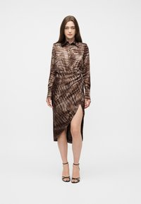 Γυναίκα με μακριά καστανά μαλλιά, φορώντας καφέ φόρεμα με κουμπιά και σχέδιο, με σκίσιμο μέχρι το μηρό και μαύρες σανδάλες με λουριά, στέκεται μπροστά σε ένα απλό φόντο.