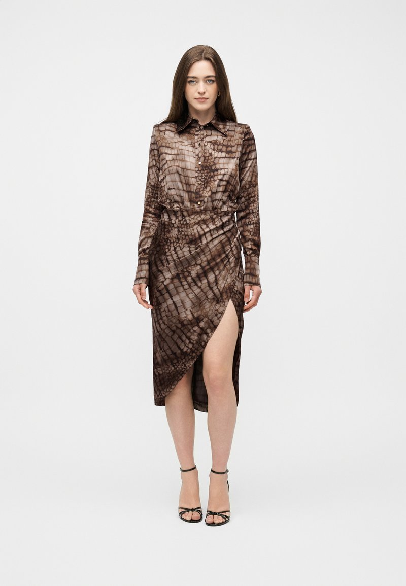 Γυναίκα με μακριά καστανά μαλλιά, φορώντας καφέ φόρεμα με κουμπιά και σχέδιο, με σκίσιμο μέχρι το μηρό και μαύρες σανδάλες με λουριά, στέκεται μπροστά σε ένα απλό φόντο.