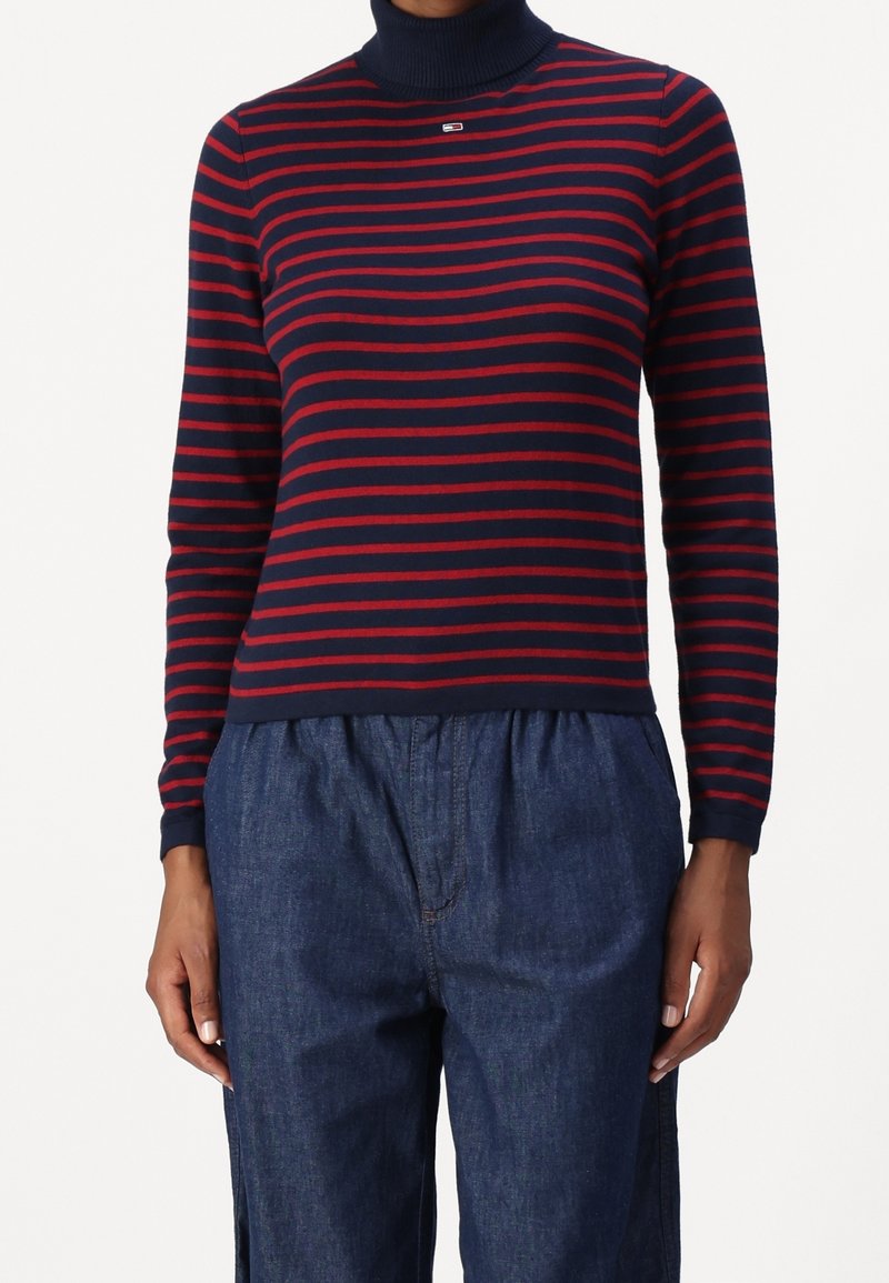 Pull à col roulé à manches longues à rayures bleu marine et rouges, fabriqué en tissu doux. Présente un petit logo sur la poitrine et une silhouette ajustée.