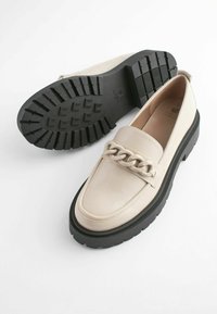 Beige nahka loaferit paksulla mustalla pohjalla, jossa on kohotettu ketjuyksityiskohta päällisessä. Pintakuvioitu pohja tarjoaa pidon.