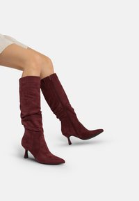 Bordeauxrode suède kniehoge laarzen met een slordig ontwerp, een puntige neus en een ritssluiting aan de achterkant, voorzien van een kleine stilettohak.