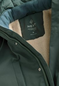 Interior verde cu un căptușeală moale din fleece bej. Prezintă un fermoar, închidere cu buton tip snap și un eticheta cu logo-ul "NEXT" și instrucțiuni de îngrijire.
