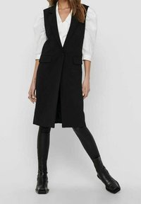 Czarna kamizelka o dopasowanym kroju z eleganckim designem, z głębokim dekoltem w serek, dwiema przednimi kieszeniami oraz zestawiona z czarnymi legginsami i botkami za kostkę.
