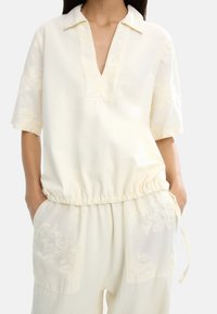 Bluzka w kolorze kremowym z krótkim rękawem, dekoltem w kształcie litery V i elastycznym dołem. Posiada kwiatowe hafty na rękawach i kieszeniach. Wykonana z miękkiego, lekkiego materiału.