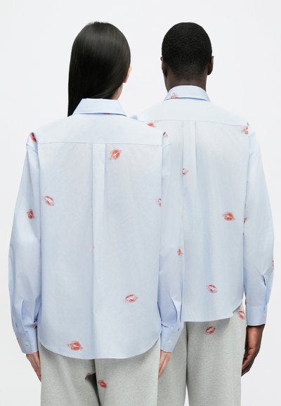 Light blue button-up shirt featuring red lipstick kiss prints, long sleeves, and a curved hem. Back design includes a center pleat.