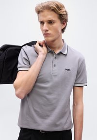 Γκρι πόλο μπλούζα με μαύρες και λευκές λεπτομέρειες, με τρεις κουμπότρυπες και λογότυπο. Το μοντέλο κρατάει ένα μαύρο μπουφάν πάνω σε έναν ώμο.