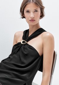 Top halter in satin nero con dettaglio nodo e un accento metallico. Texture liscia e design fluido, caratterizzato da una drappeggiatura sottile.