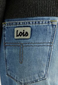 Niebieskie dżinsy z widoczną szaro skórzaną naszywką z napisem „Lois” na tylnej kieszeni. Materiał ma gładką teksturę i żółte przeszycia.