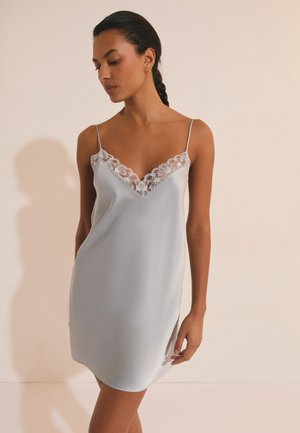 Femme aux cheveux tressés portant une robe nuisette en satin bleu clair avec bordure en dentelle, debout contre un fond beige, regardant vers le bas.