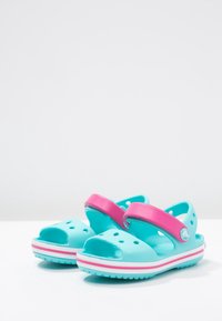Lasten turkoosit sandaalit vaaleanpunaisilla säädettävillä remmeillä ja ilmanvaihtorei'illä, esillä pelkällä valkoisella taustalla.