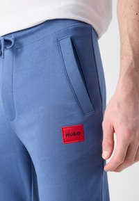 Gaiši zili sporta bikses, kas izgatavotas no kokvilnas maisījuma, ar sānu kabatām un sarkanu kvadrātveida uzlīmi, kurā melnā krāsā ir uzraksts "HUGO".