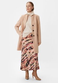 Beżowy płaszcz na kremowym golfie, zestawiony z długą, wzorzystą spódnicą w różowym, czarnym i brązowym kolorze. Beżowe, wysokie buty na obcasie dopełniają stylizację.
