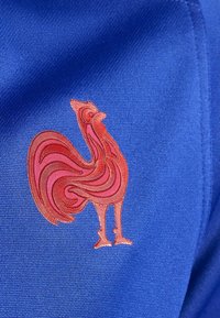 Blått sporttyg har en röd broderad tupp-logotyp med intrikata snirkliga linjer och detaljer på bröstområdet.