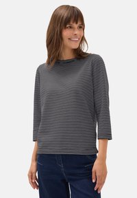 Kvinna som ler, iförd en svart och vit horisontellt randig långärmad tröja och mörkblå jeans, stående mot en enfärgad bakgrund.