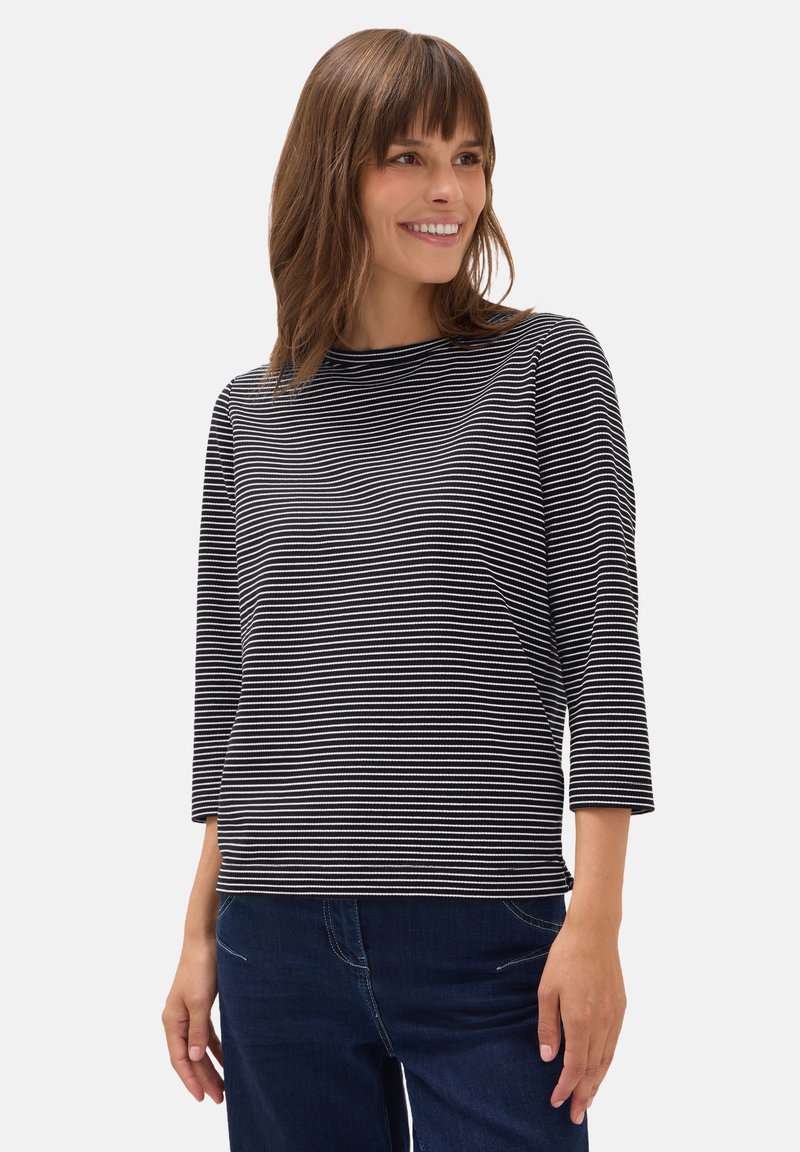 Kvinna som ler, iförd en svart och vit horisontellt randig långärmad tröja och mörkblå jeans, stående mot en enfärgad bakgrund.