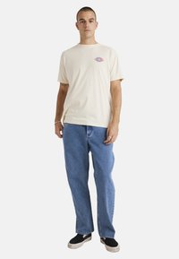 Camiseta de algodón color crema con un gráfico de un ojo rojo y azul en el lado izquierdo del pecho, combinada con pantalones vaqueros azules y zapatos negros sin cordones.