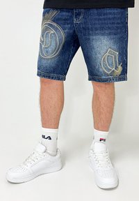 Denim shorts in dark blue with gold embroidered patterns. Paired with white sneakers and socks featuring brand logo. Casual fit.