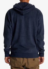 Sudadera con capucha de forro polar azul marino con un acabado texturizado, capucha con cordón y puños acanalados, combinada con pantalones cargo beige con bolsillos traseros.