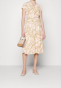 Vestido rosa claro con patrón de cadenas doradas, mangas cortas, cierre de botones en la parte delantera y cinturón en la cintura. Combinado con un bolso bicolor y tacones nude.